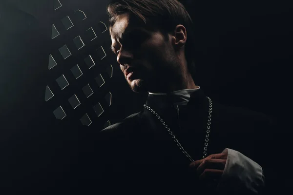 Young serious catholic priest touching cross on his necklace in dark near confessional grille — Stock Photo