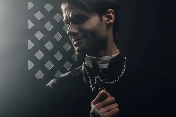 Young worried catholic priest touching cross on his necklace in dark near confessional grille — Stock Photo