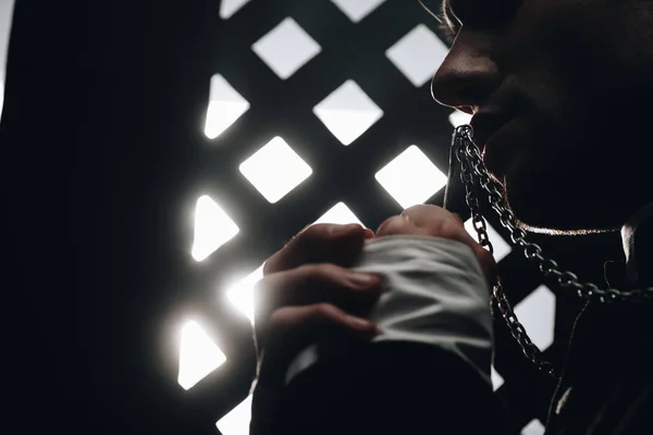Visão cortada do padre católico beijando cruz em seu colar perto da grade confessional no escuro com raios de luz — Fotografia de Stock