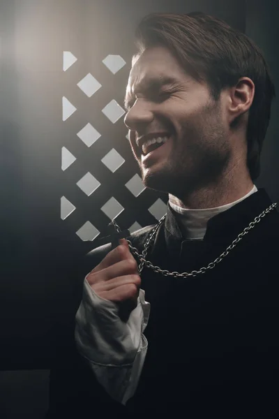 Sarcástico sacerdote católico riendo mientras toca la cruz en su collar cerca de la rejilla confesional en la oscuridad con rayos de luz - foto de stock
