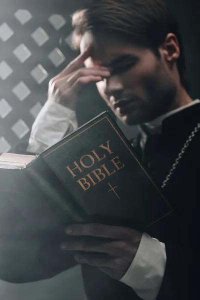 Joven sacerdote católico serio tocando la cara mientras lee la biblia cerca de la rejilla confesional en la oscuridad con rayos de luz - foto de stock