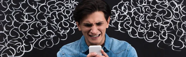 Tiro panorâmico de homem jovem preocupado usando smartphone, com vapor de orelhas desenhando em quadro-negro — Fotografia de Stock