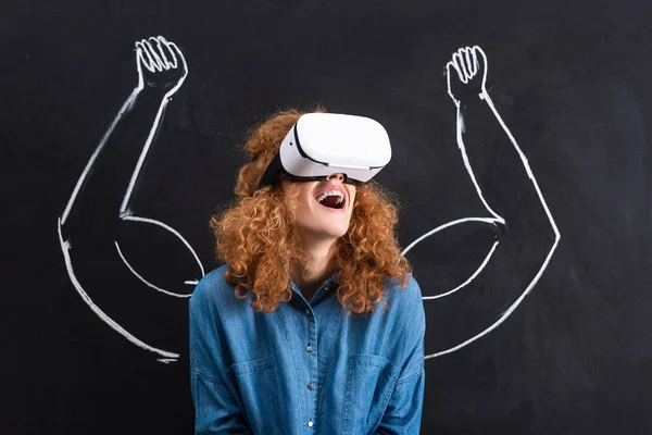 Excited girl using virtual reality headset with strong arms drawing on blackboard — Stock Photo