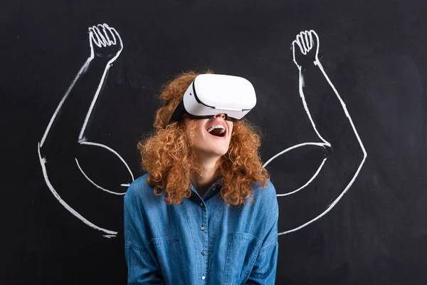 Chica excitada usando auriculares de realidad virtual con fuertes brazos dibujando en pizarra - foto de stock