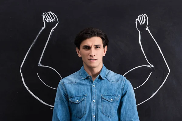 Pensive young man with strong arms drawing on blackboard — Stock Photo