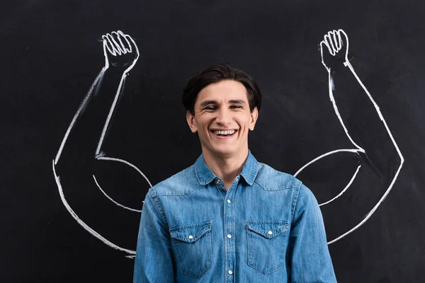 Handsome smiling man with strong arms drawing on blackboard — Stock Photo