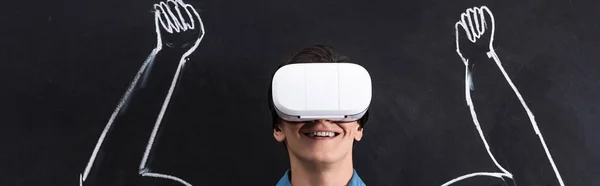 Tiro panorâmico de jovem sorridente usando fone de ouvido realidade virtual, com braços musculares desenhando em quadro-negro — Fotografia de Stock