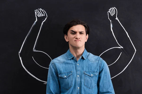 Skeptical young man with strong arms drawing on blackboard — Stock Photo