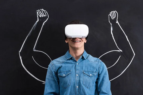Happy young man using virtual reality headset, with muscular arms drawing on blackboard — Stock Photo