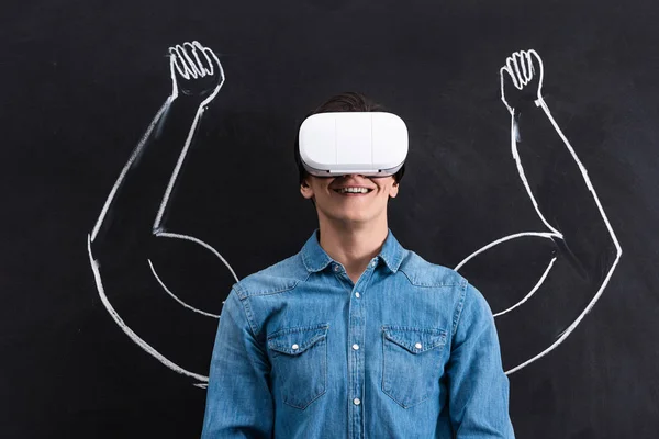 Joven feliz usando auriculares de realidad virtual, con brazos musculares dibujando en pizarra - foto de stock