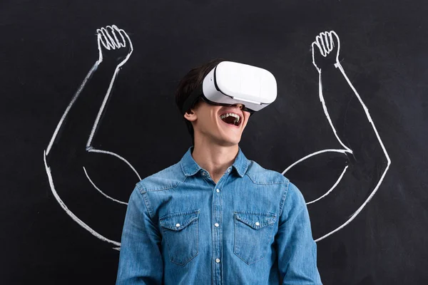 Excited young man using virtual reality headset, with muscular arms drawing on chalkboard — Stock Photo