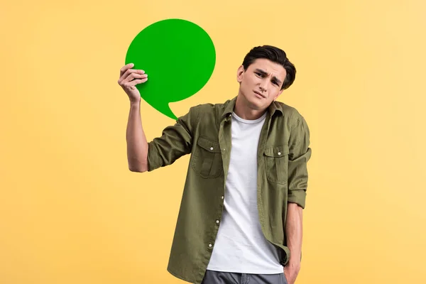Hombre confundido sosteniendo burbuja de habla verde, aislado en amarillo - foto de stock