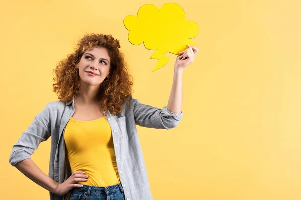 Mujer sonriente pensativa sosteniendo la burbuja del habla vacía en forma de nube, aislada en amarillo - foto de stock