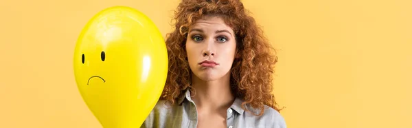 Panoramic shot of upset redhead woman holding balloon with sad face, isolated on yellow — Stock Photo