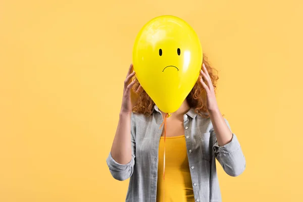 Mujer joven sosteniendo triste globo en frente de la cara, aislado en amarillo - foto de stock