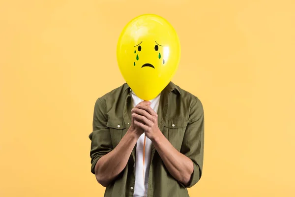Young man holding crying balloon in front of face, isolated on yellow — Stock Photo