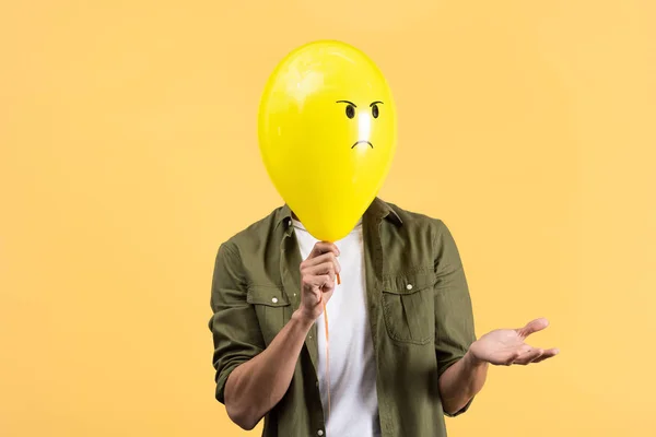 Giovane uomo con gesto di scrollata di spalle tenendo palloncino arrabbiato davanti al viso, isolato su giallo — Foto stock