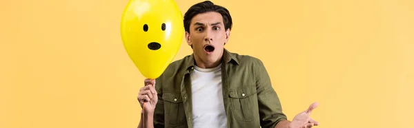 Disparo panorámico de un joven asustado sosteniendo un globo impactado, aislado en amarillo - foto de stock