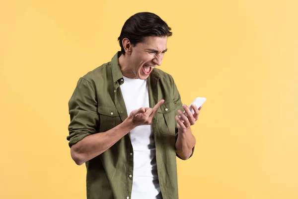 Joven agresivo gritando y mostrando el dedo medio al teléfono inteligente, aislado en amarillo - foto de stock