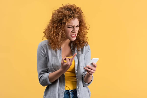 Mujer pelirroja estresada mostrando el dedo medio al teléfono inteligente, aislado en amarillo - foto de stock