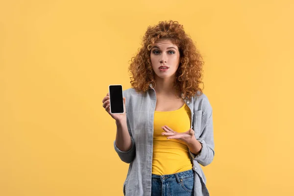 Mujer pelirroja confundida mostrando teléfono inteligente con pantalla en blanco, aislado en amarillo - foto de stock