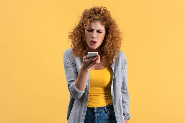 Aggressive redhead woman yelling on smartphone, isolated on yellow — Stock Photo