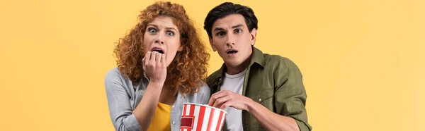 Panoramic shot of frightened couple watching movie with bucket of popcorn, isolated on yellow — Stock Photo