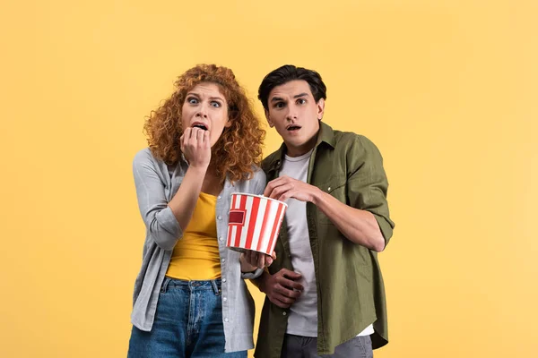 Pareja asustada viendo película de terror con cubo de palomitas de maíz, aislado en amarillo - foto de stock