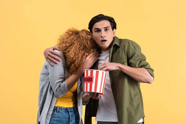 Casal assustado assistindo filme com balde de pipoca, isolado em amarelo — Fotografia de Stock