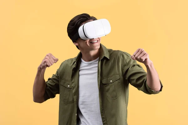 Jovem feliz segurando punhos e jogando fone de ouvido realidade virtual, isolado no amarelo — Fotografia de Stock