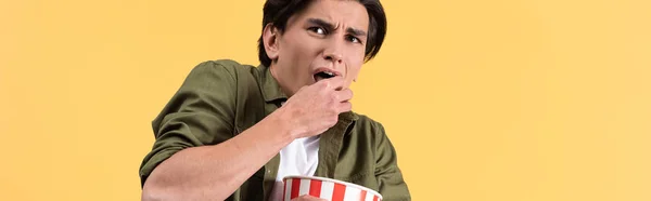 Plano panorámico de un joven asustado viendo películas de terror y comiendo palomitas de maíz, aislado en amarillo - foto de stock