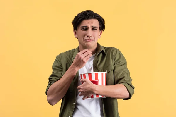 Засмучений молодий чоловік дивиться фільм з відром попкорну, ізольований на жовтому — стокове фото