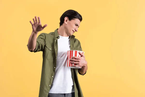 Irritated young man watching movie with bucket of popcorn, isolated on yellow — Stock Photo