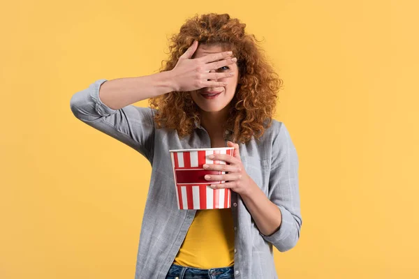 Chica asustada cerrando los ojos mientras mira película de terror con cubo de palomitas de maíz, aislado en amarillo — Stock Photo