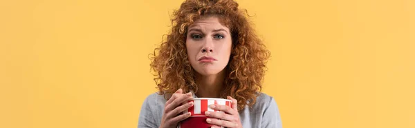 Panoramic shot of sad girl watching movie with bucket of popcorn, isolated on yellow — Stock Photo