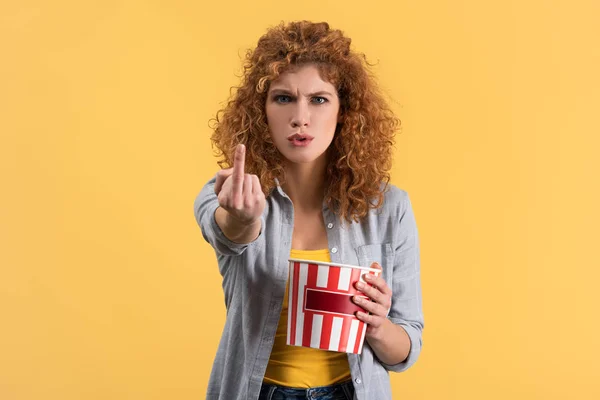 Aggressive girl showing middle finger while watching movie with bucket of popcorn, isolated on yellow — Stock Photo