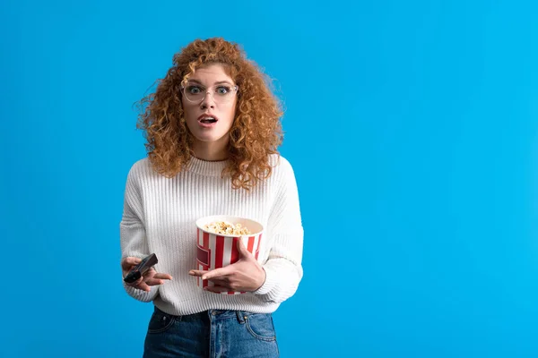 Shocked girl watching tv with remote control and bucket of popcorn, isolated on blue — Stock Photo