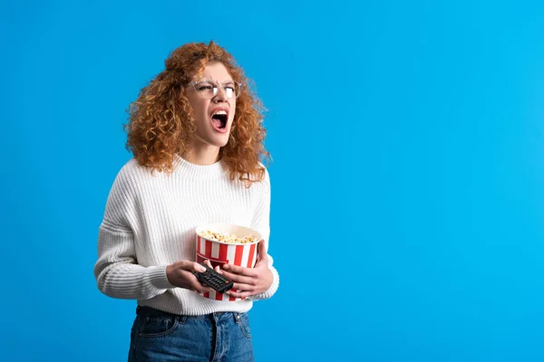 Wütende Mädchen schreien und fernsehen mit Fernbedienung und Eimer Popcorn, isoliert auf blau — Stockfoto