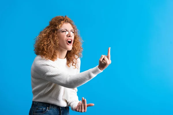 Irritado gritando ruiva menina em óculos mostrando dedo médio, isolado em azul — Fotografia de Stock