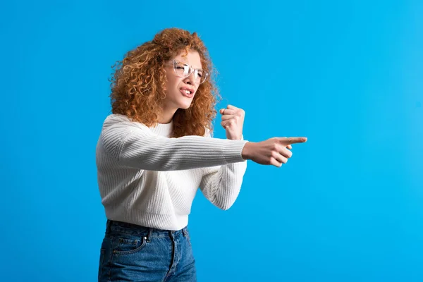 Aggressiva rossa ragazza in occhiali puntando e tenendo il pugno, isolata su blu — Foto stock