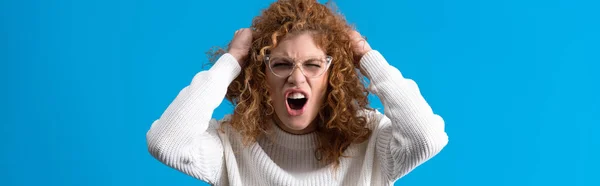 Panoramic shot of aggressive screaming redhead girl in eyeglasses, isolated on blue — Stock Photo
