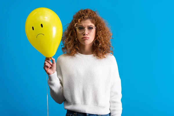 Sad girl holding yellow balloon with upset face, isolated on blue — Stock Photo