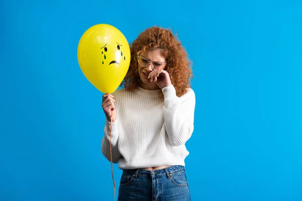 Lonely redhead girl holding yellow balloon with sad face, isolated on blue — Stock Photo