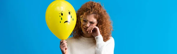 Panoramic shot of upset redhead girl holding yellow balloon with crying face, isolated on blue — Stock Photo