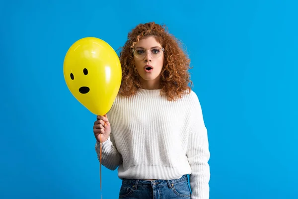 Shocked girl holding yellow balloon with surprised face, isolated on blue — Stock Photo