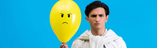 Disparo panorámico de joven agresivo sosteniendo globo enojado amarillo, aislado en azul - foto de stock