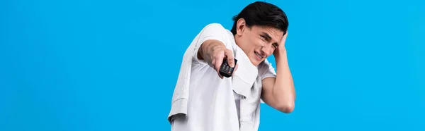 Panoramic shot of frightened man watching tv with remote controller, isolated on blue — Stock Photo