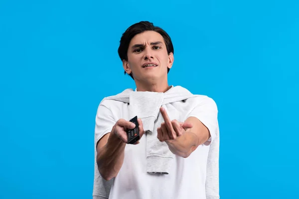 Aggressive man showing middle finger while watching tv with remote controller, isolated on blue — Stock Photo