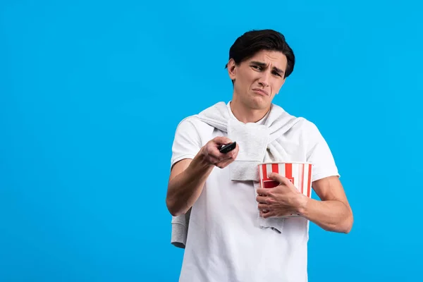 Jovem triste assistindo tv com controle remoto e pipoca, isolado em azul — Fotografia de Stock