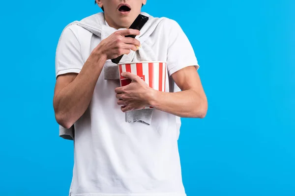 Обрізаний вид шокованого чоловіка, який дивиться телевізор з пультом дистанційного керування і попкорном, ізольований на синьому — стокове фото